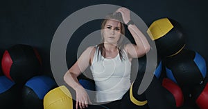 Portrait of beautiful young Caucasian athlete woman sitting on gym floor among cross fit balls, exhausted after workout.