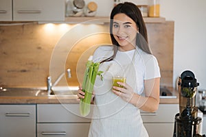 Portrait of beautiful and young female in apron standing in the kitchen. Woman hold glass with green healthy beverage in