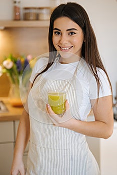 Portrait of beautiful and young female in apron standing in the kitchen. Woman hold glass of green healthy beverage