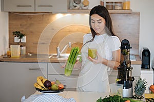 Portrait of beautiful and young female in apron standing in the kitchen. Woman hold glass with green healthy beverage in