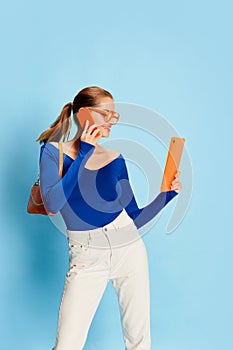 Portrait of beautiful young girl in official stylish clothes talking on phone and looking on tablet over blue background