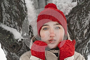 Portrait of beautiful young girl in red knitted hat and mittens warms up near tree in park. Sad girl. Pleasant winter weather