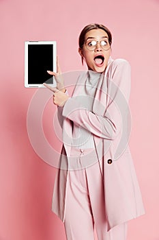 Portrait of beautiful young girl in suit and glasses posing, emotionally pointing on tablet over pink background. Good