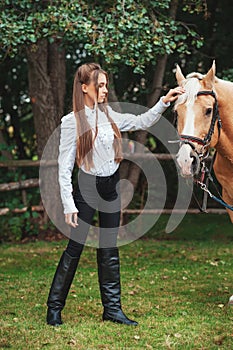 Portrait beautiful young girl in white shirt and black pants with beauty long hair next horse in forest. Fashionable elegance woma