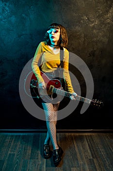 Portrait of beautiful young hipster woman with curly hair with red guitar in neon lights. Rock musician is playing