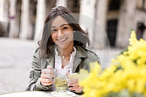 Portrait of beautiful young hispanic woman drinking a cup of camomile tea in cafeteria with mimosa flower bouquet on the table