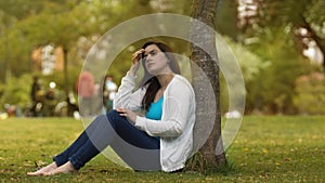 Portrait of beautiful young Hispanic woman sitting under a tree in the middle of a park with a pensive attitude against a