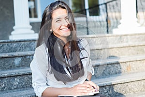 Portrait of a beautiful young Indian girl, business woman, smiling, sitting on the steps of an office building, holding a notepad