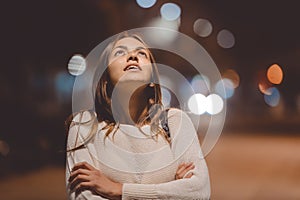 Portrait of beautiful young lady looking up, city street in the night, evening lights bokeh background outdoors