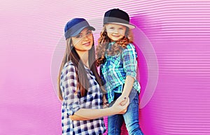 Portrait of beautiful young mother with little girl child wearing checkered shirts, baseball cap in city on pink background