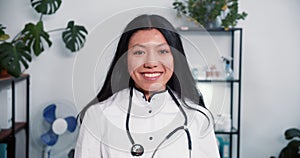 Portrait of beautiful young multiethnic doctor woman wearing white lab coat, stethoscope smile at camera at light office