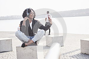 Portrait of a beautiful young pretty African American girl sitting on the beach or lake and listening to music in her