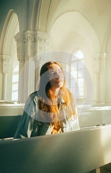 Portrait of a beautiful young red-haired woman sitting on a bench in an old church, illuminated by divine light. Faith and