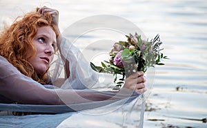 Portrait of beautiful young sexy red-haired girl with a longing, Lady of Shalott, with a bouquet of flowers in a boat, copy space