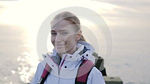 Portrait of beautiful young smiling woman in jacket with backpack on pier with sea on a background