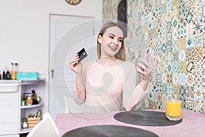 Portrait of a beautiful young student girl sitting indoors using laptop computer chatting by mobile phone holding credit card