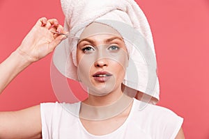 Portrait of beautiful young woman 20s wrapped in white towel after shower plucking her eyebrows with tweezer