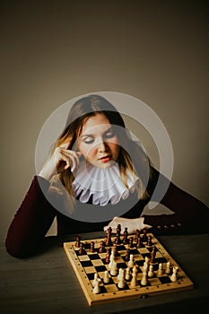 Portrait of a beautiful young woman in an antique dress with a white frill, who is sitting in front of a chess board and thinking