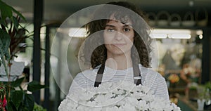 Portrait of beautiful young woman in apron standing in flower shop holding bouquet and smiling looking at camera