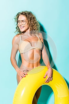 Portrait of beautiful young woman in bikini and glasses playing with inflatable yellow float isolated on blue background