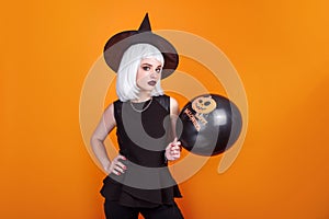 Portrait of beautiful young woman in black witch halloween costume with black balloon over orange background