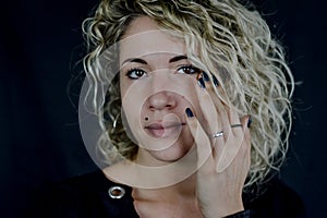 Portrait of a beautiful young woman with blond curly hair who fixes her eye makeup with her finger