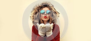 Portrait of beautiful young woman blowing her lips with red lipstick sending sweet air kiss wearing jacket with fur hood on blank