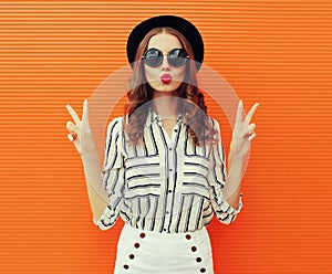 Portrait beautiful young woman blowing her lips sending sweet air kiss wearing a white striped shirt, black round hat on orange