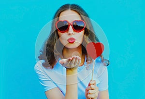 Portrait of beautiful young woman blowing red lips sweet air kiss with red heart shaped lollipop wearing sunglasses