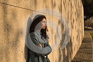 Portrait of beautiful young woman, brunette, with curly hair, wearing jacket with arms crossed leaning against a wall with pensive