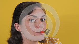 Portrait of beautiful young woman cheerfully blowing party horn and happy birthday hat on yellow background slow motion