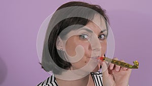 Portrait of a beautiful young woman cheerfully blowing a party horn on a purple background slow motion