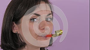 Portrait of a beautiful young woman cheerfully blowing a party horn on a purple background slow motion