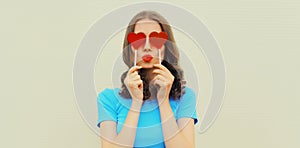 Portrait of beautiful young woman covering her eyes with red heart shaped lollipop blowing her lips with lipstick sending sweet