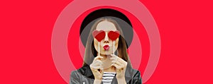 Portrait of beautiful young woman covering her eyes with red heart shaped lollipop blowing lipstick sending sweet air kiss on