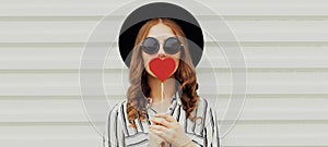 Portrait beautiful young woman covering her mouth with red heart shaped lollipop on a white background