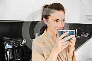 Portrait of beautiful young woman, drinking coffee, freshly made cappuccino, smiling pleased, enjoying her morning