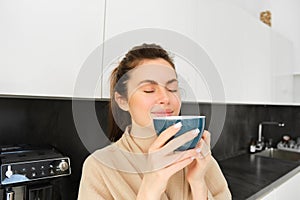 Portrait of beautiful young woman, drinking coffee, freshly made cappuccino, smiling pleased, enjoying her morning