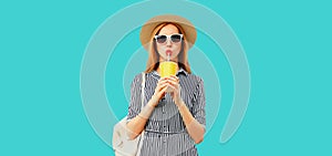 Portrait of beautiful young woman drinking fruit juice wearing summer straw round hat, striped dress  on blue background