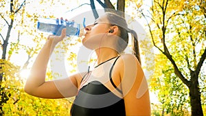 Portrait of beautiful young woman drinking water after doing sports and fitness exercises in autumn park.