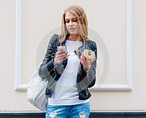 Portrait of a beautiful young woman eating a donut, looks at her smart phone on the street European city. Outdoor.