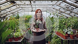 Portrait of beautiful young woman farmer in apron standing inside greenhouse, holding pot plant and smiling. Orcharding