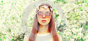 Portrait of beautiful young woman and flowers wearing floral headband, sunglasses on spring blooming garden background