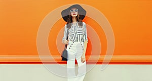 Portrait of beautiful young woman with handbag wearing white striped shirt, black round hat on orange background