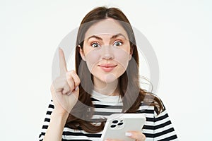 Portrait of beautiful young woman, has an idea, suggestion, shows one finger and holds smartphone, points up, white