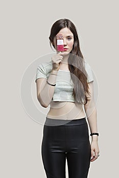 Portrait of beautiful young woman holding ice cream bar over eye against gray background