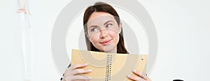 Portrait of beautiful young woman, holding daily planner, writing in her diary, making notes in notebook and smiling