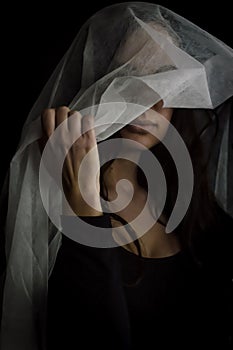 Portrait of a beautiful young woman holding a transparent white fabric covering her head and face leaving only her mouth visible