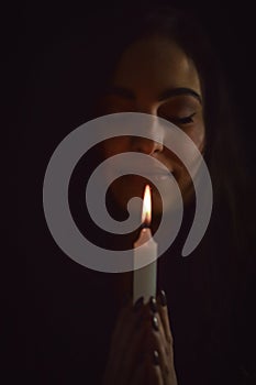 Portrait of a beautiful young woman with long hair holding a lit candle in her hand and praying with closed eyes