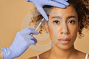 Portrait of beautiful young woman looking at camera while getting face injections  over beige background